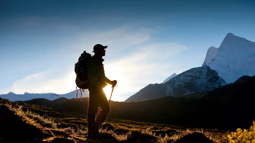 Silhouetted hiker with a backpack and trekking poles stands on a mountain trail at sunrise. Majestic snow-capped peaks are visible against the clear sky.