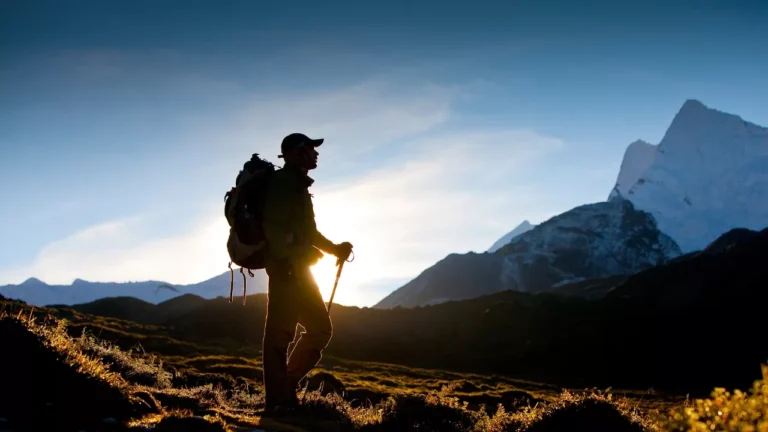 Silhouetted hiker with a backpack and trekking poles stands on a mountain trail at sunrise. Majestic snow-capped peaks are visible against the clear sky.