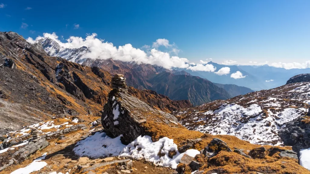Mountain landscape with rugged peaks and scattered snow patches under a clear blue sky. A cairn stands on a rocky foreground, evoking serenity.