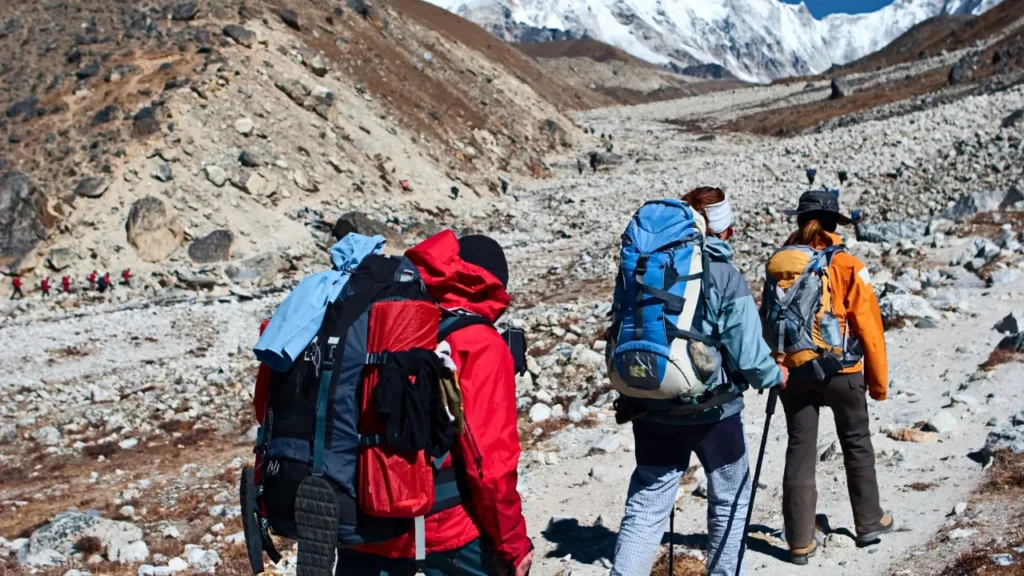 A group of hikers with backpacks trek through a rocky, mountainous landscape. Snow-capped peaks are visible in the distance, conveying a sense of adventure.