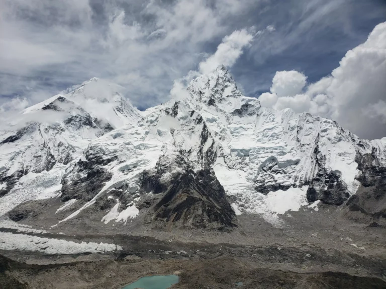 Snow-covered mountain peaks under a partly cloudy sky, with rugged terrain and a small turquoise lake at the base, conveying majesty and tranquility