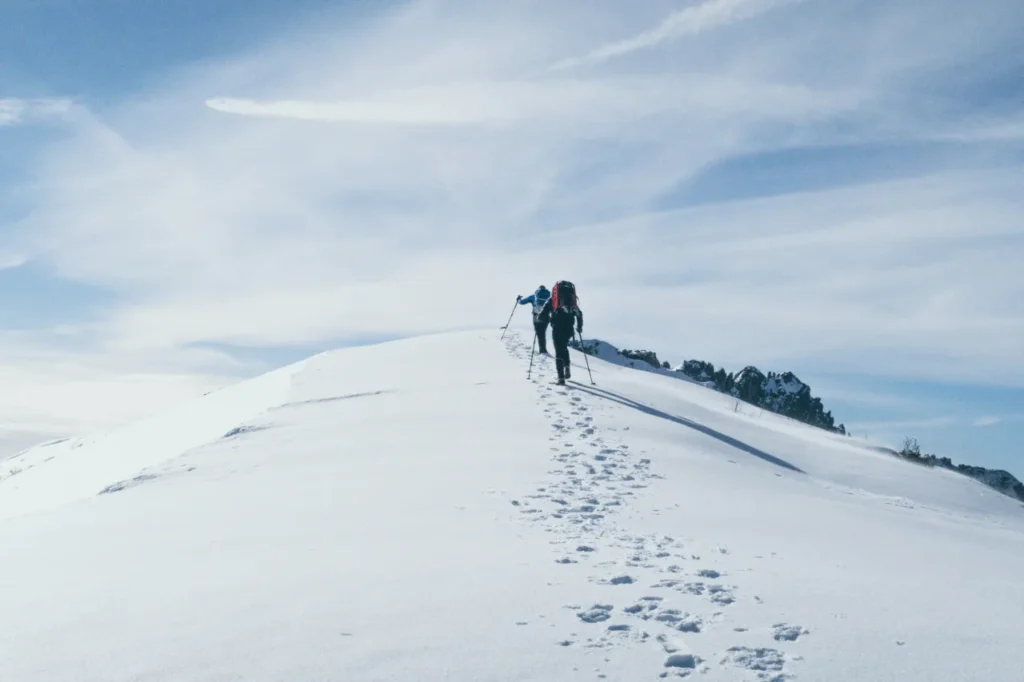 Two mountaineers with backpacks and trekking poles ascend a steep, snow-covered mountain ridge towards a crest.