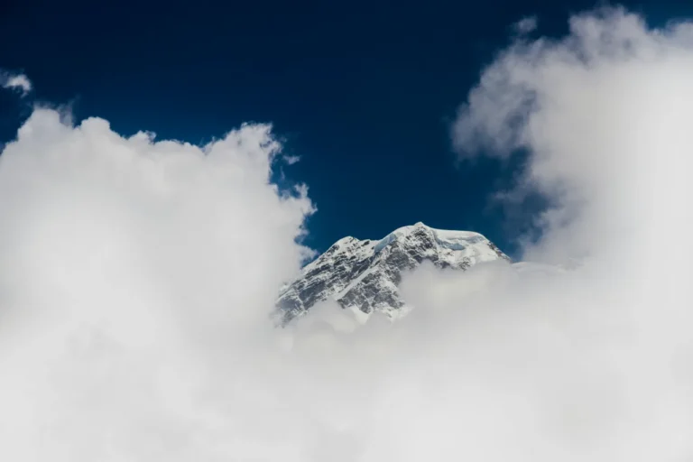 A majestic snow-capped mountain peak poking through a dense layer of fluffy white clouds under a dark, deep blue sky.
