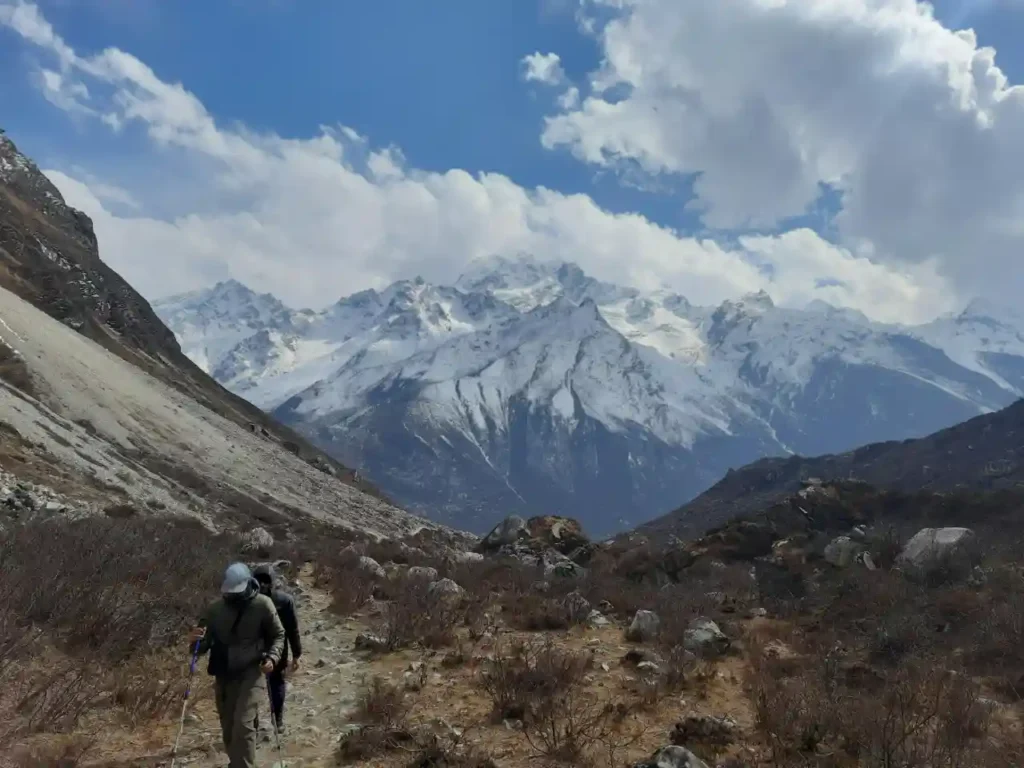 Two hikers with trekking poles walk along a rocky path in a high-altitude mountain valley, with a backdrop of massive, jagged, snow-capped mountain peaks under a blue sky with white clouds.