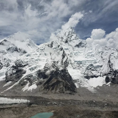 Snow-covered mountain peaks under a partly cloudy sky, with rugged terrain and a small turquoise lake at the base, conveying majesty and tranquility