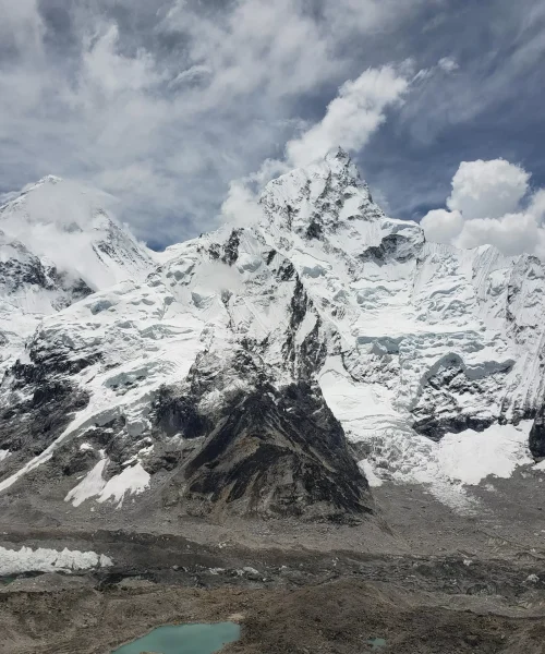 Snow-covered mountain peaks under a partly cloudy sky, with rugged terrain and a small turquoise lake at the base, conveying majesty and tranquility