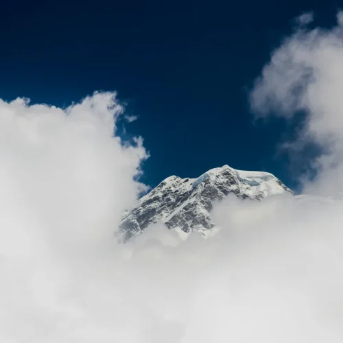 A majestic snow-capped mountain peak poking through a dense layer of fluffy white clouds under a dark, deep blue sky.
