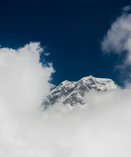 A majestic snow-capped mountain peak poking through a dense layer of fluffy white clouds under a dark, deep blue sky.